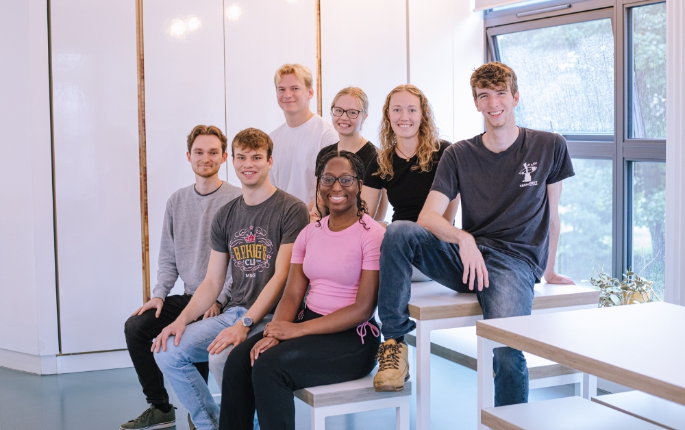 Image: 7 of the interns at Crux Product Design, sat on a table in the office with a white background